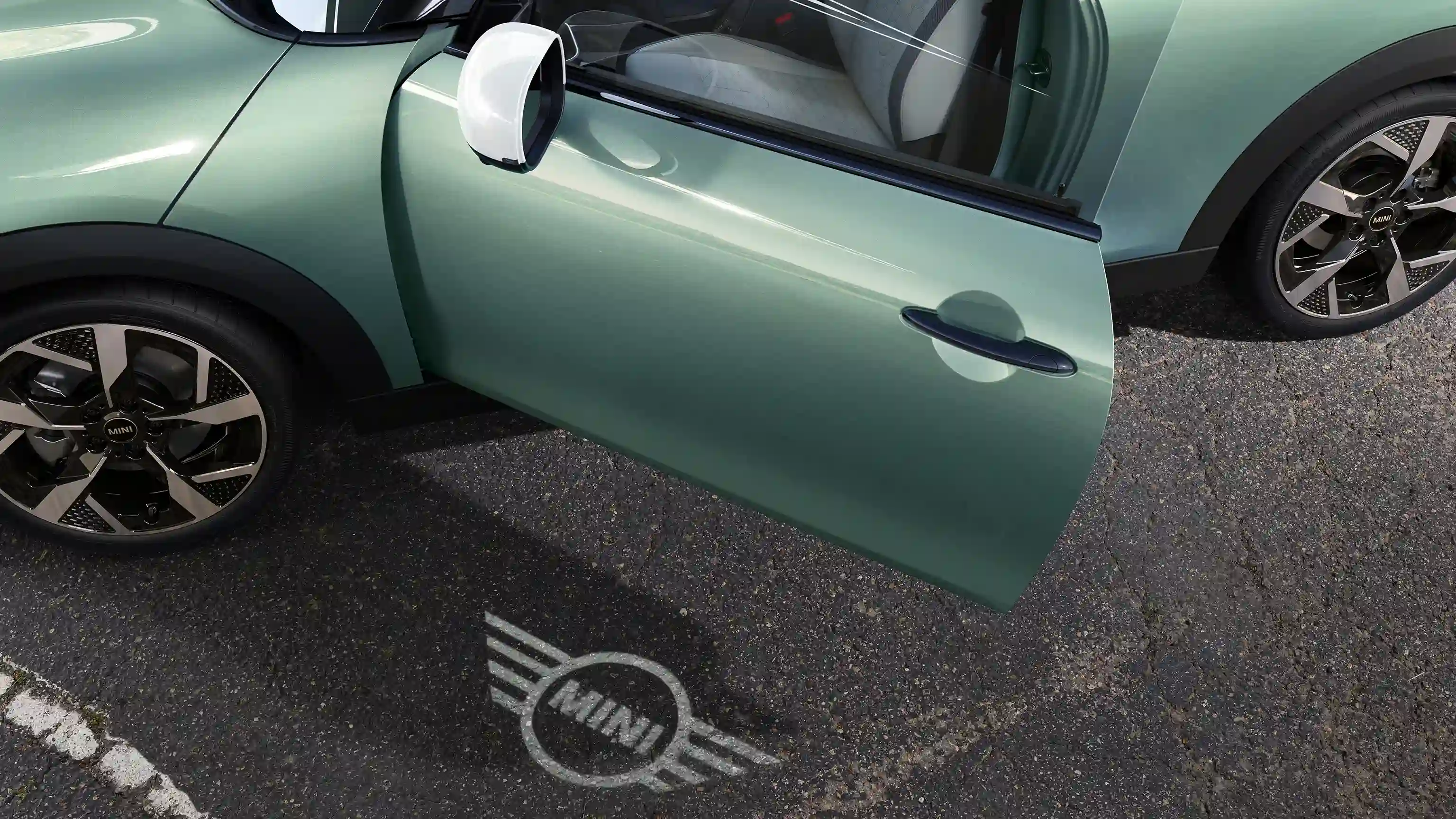 Pogled od zunaj na MINI Cooper z odprtimi vrati in pozdravno lučjo z logotipom MINI, projiciranim na tla 