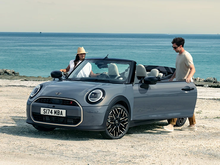 Levi pogled od spredaj na MINI Cooper Cabrio v barvi Copper Grey na plaži z dvema osebama ob njem.