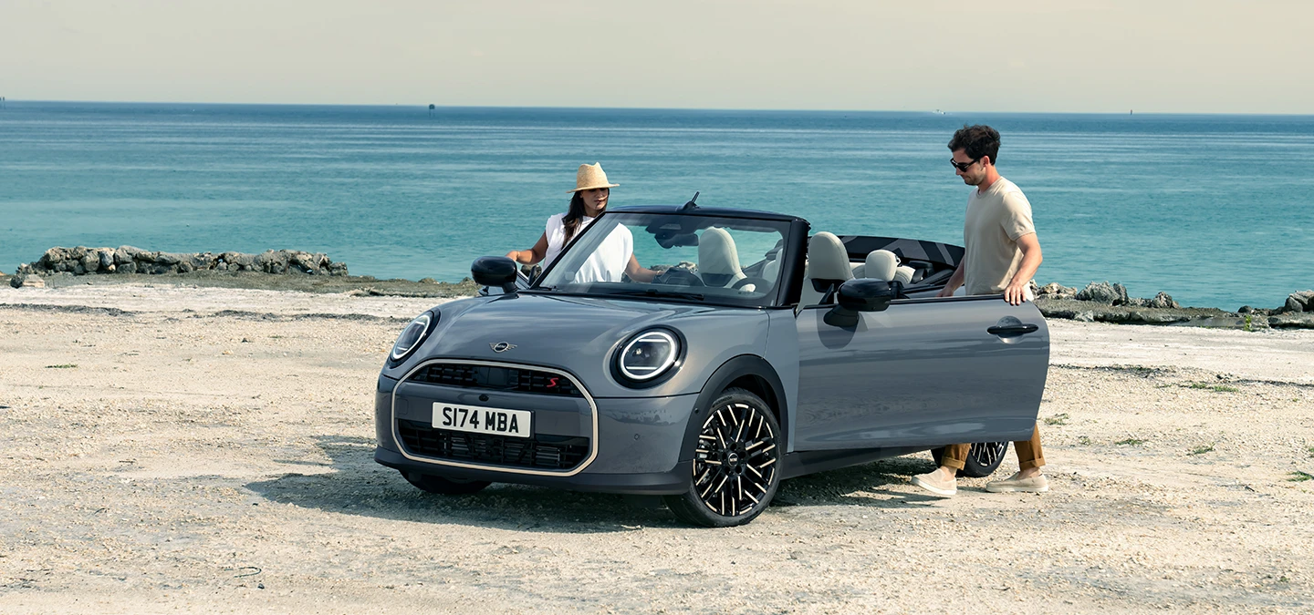 Levi pogled od spredaj na MINI Cooper Cabrio v barvi Copper Grey na plaži z dvema osebama ob njem.