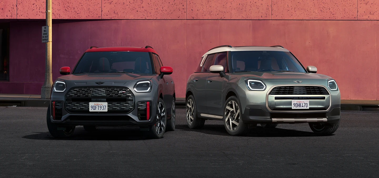 Pogled od spredaj na MINI Countryman JCW v barvi Legend Grey z rdečo streho ter na MINI Countryman v barvi Smokey Green z izrazito streho Vibrant Silver, parkirana drug ob drugem.