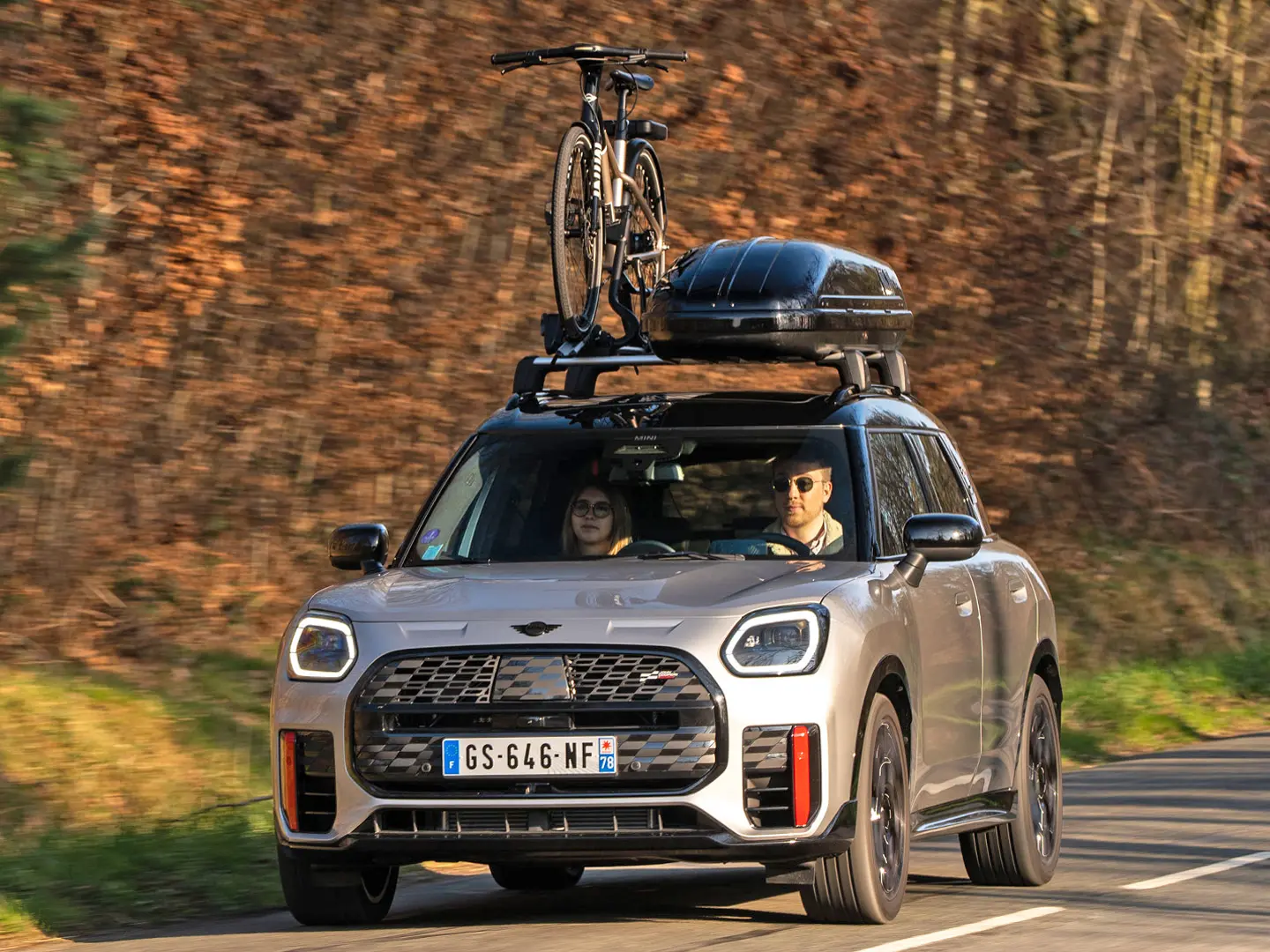 MINI Countryman Cooper v srebrni barvi na gorski cesti, opremljen s strešnimi nosilci MINI, strešnim kovčkom MINI, strešnim nosilcem za kolo MINI z enim nameščenim kolesom in krilcem MINI John Cooper Works – pogled od spredaj. 