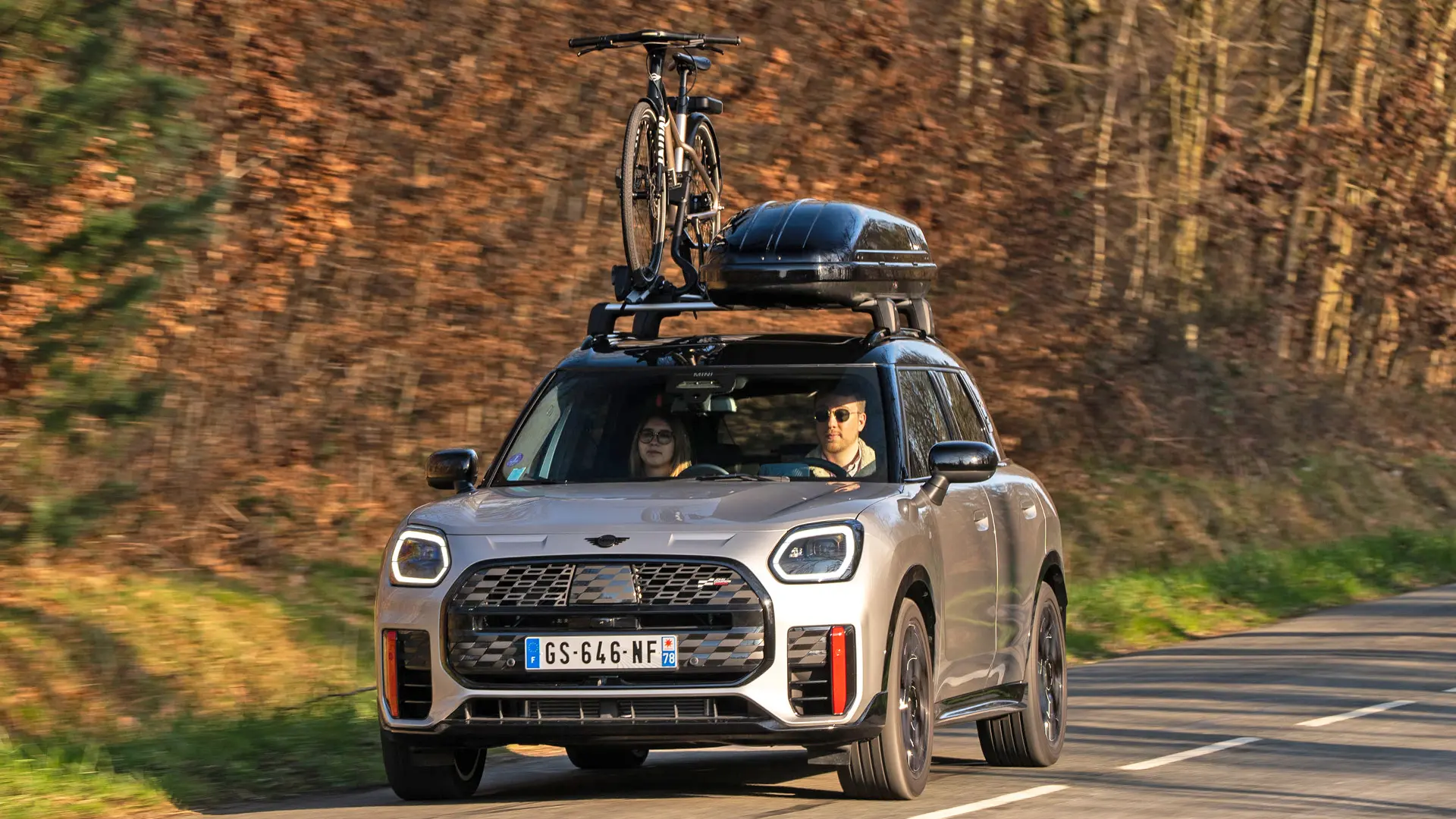 MINI Countryman Cooper v srebrni barvi na gorski cesti, opremljen s strešnimi nosilci MINI, strešnim kovčkom MINI, strešnim nosilcem za kolo MINI z enim nameščenim kolesom in krilcem MINI John Cooper Works – pogled od spredaj.