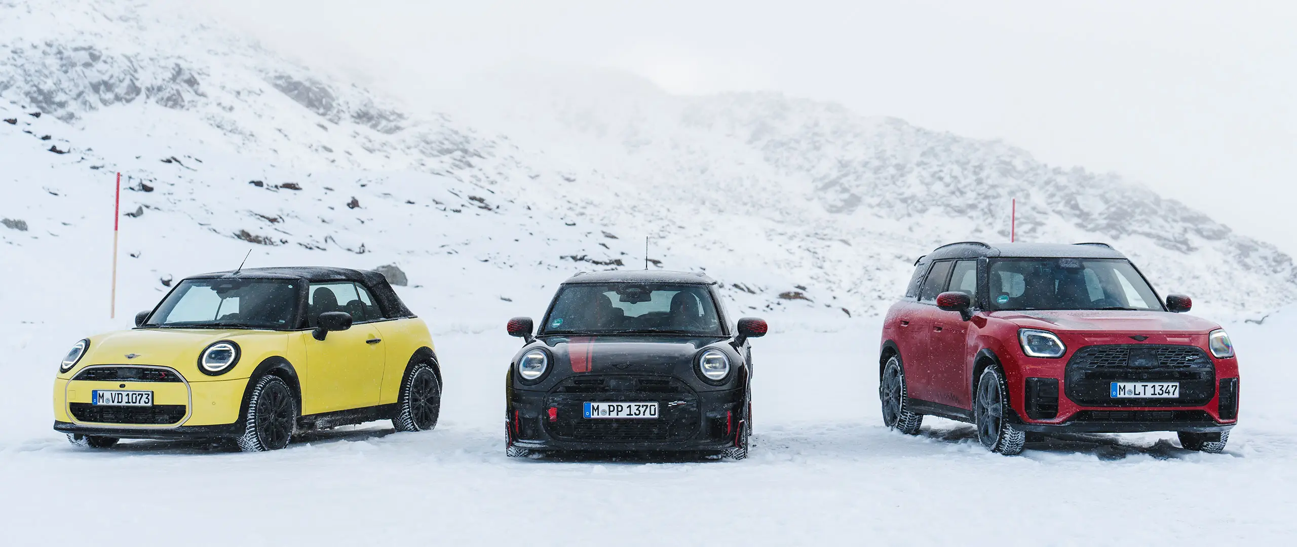 MINI Cooper Cabrio v barvi Sunny Side Yellow, MINI John Cooper Works v barvi Midnight Black II in MINI Countryman Cooper v barvi Chilli Red – parkiran na zasneženi gorski cesti, pogled od spredaj.