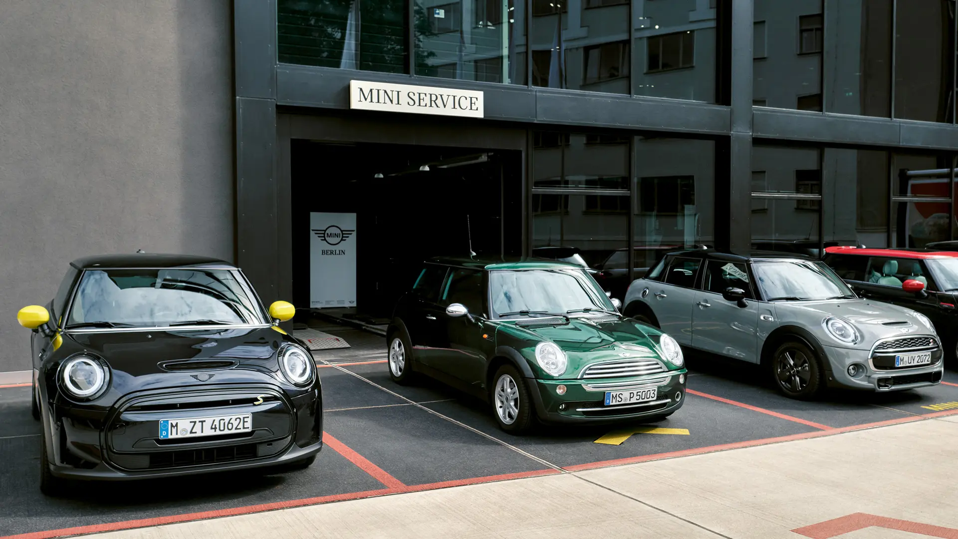 MINI Cooper Electric S v črni barvi, parkiran pred prodajnim salonom MINI, ob njem MINI Cooper 3-vratni v zeleni barvi, MINI 5-vratni v srebrni barvi in MINI JCW v črni barvi, pogled od spredaj 