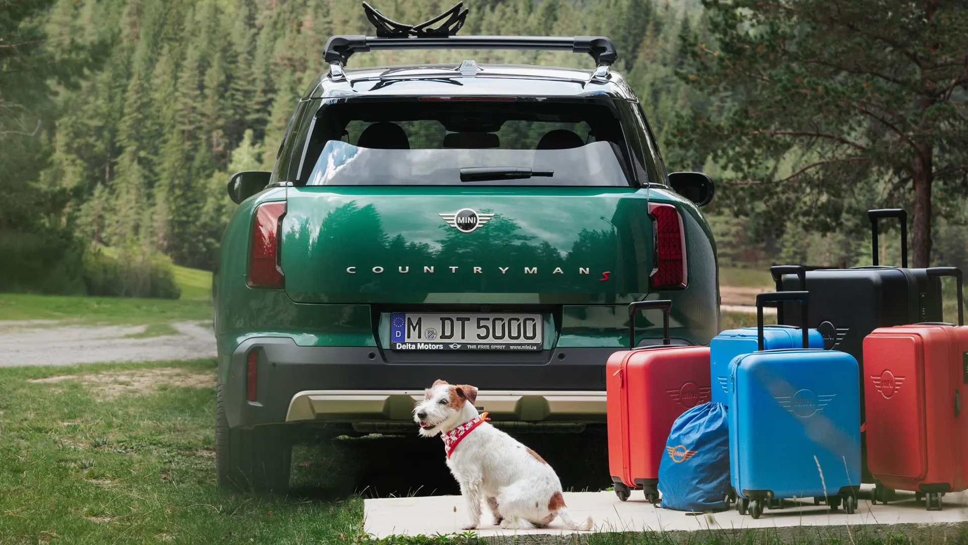MINI Countryman v zeleni barvi z različnimi kosi prtljage, na voljo v trgovini MINI Lifestyle, postavljeni za vozilom skupaj s psom pasme terier. 