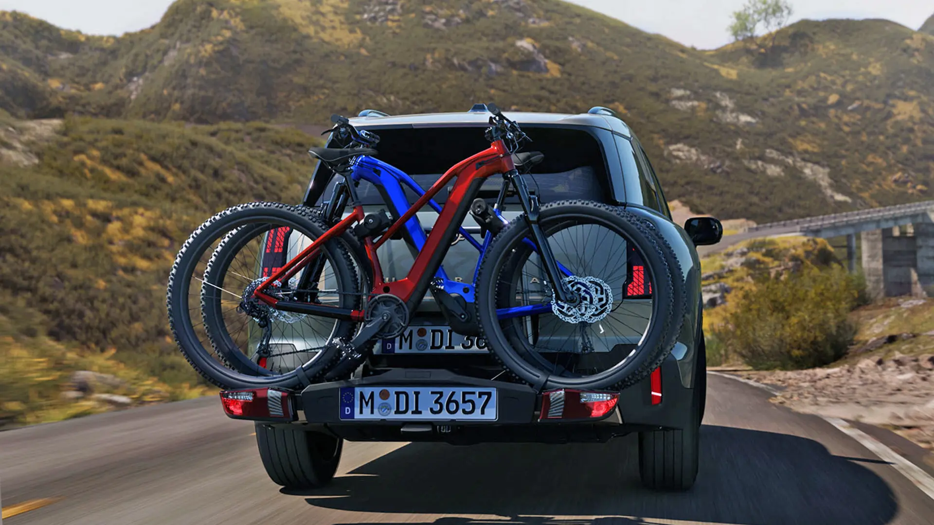 MINI Countryman v barvi Smokey Green z zadnjim nosilcem za kolesa in dvema pritrjenima gorskima kolesoma, pogled od zadaj, vožnja po gorski cesti.