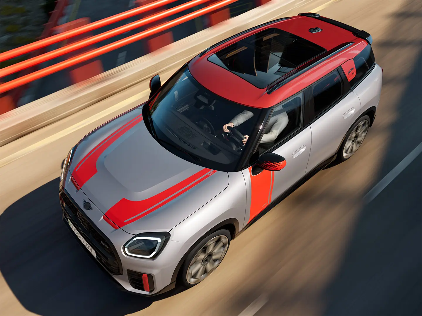 MINI Countryman John Cooper Works v barvi Melting Silver s pokrovi zunanjih ogledal, nalepkami na straneh in pokrovu motorja, streho in krilci v barvi Chili Red med vožnjo po mestni ulici – pogled od spredaj.