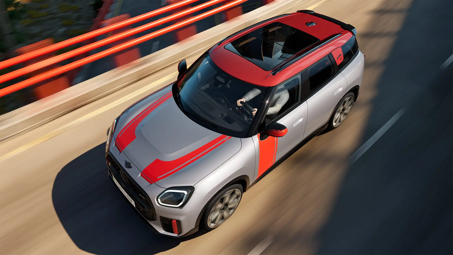 MINI Countryman John Cooper Works v barvi Melting Silver s pokrovi zunanjih ogledal, nalepkami na straneh in pokrovu motorja, streho in krilci v barvi Chili Red med vožnjo po mestni ulici – pogled od spredaj.