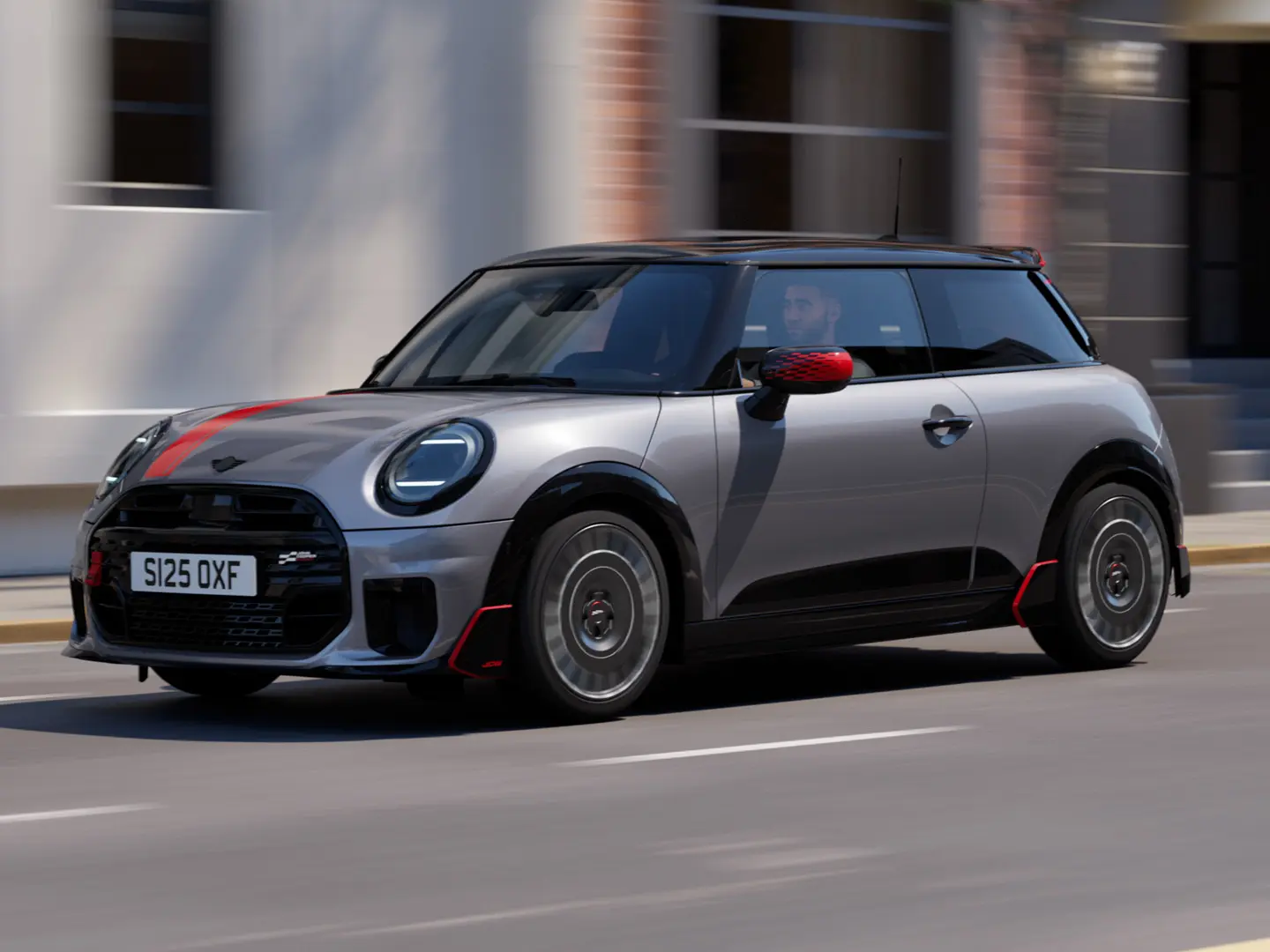MINI Cooper s 3 vrati v barvi Melting Silver z deli John Cooper Works: pokrovi zunanjih ogledal in nalepko na pokrovu motorja v barvi Chili Red, strešnim spojlerjem in krilcem v črni barvi med vožnjo po mestni ulici – pogled od spredaj.