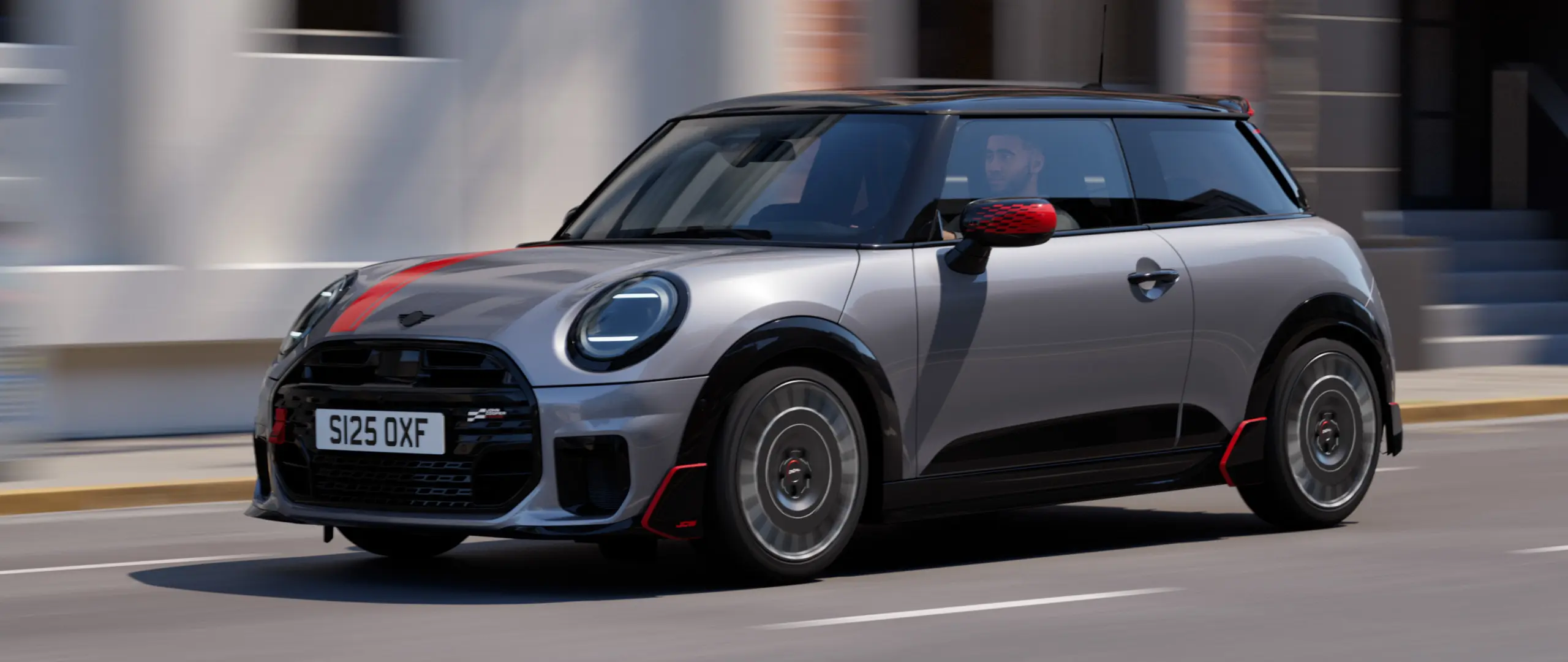 MINI Cooper s 3 vrati v barvi Melting Silver z deli John Cooper Works: pokrovi zunanjih ogledal in nalepko na pokrovu motorja v barvi Chili Red, strešnim spojlerjem in krilcem v črni barvi med vožnjo po mestni ulici – pogled od spredaj.