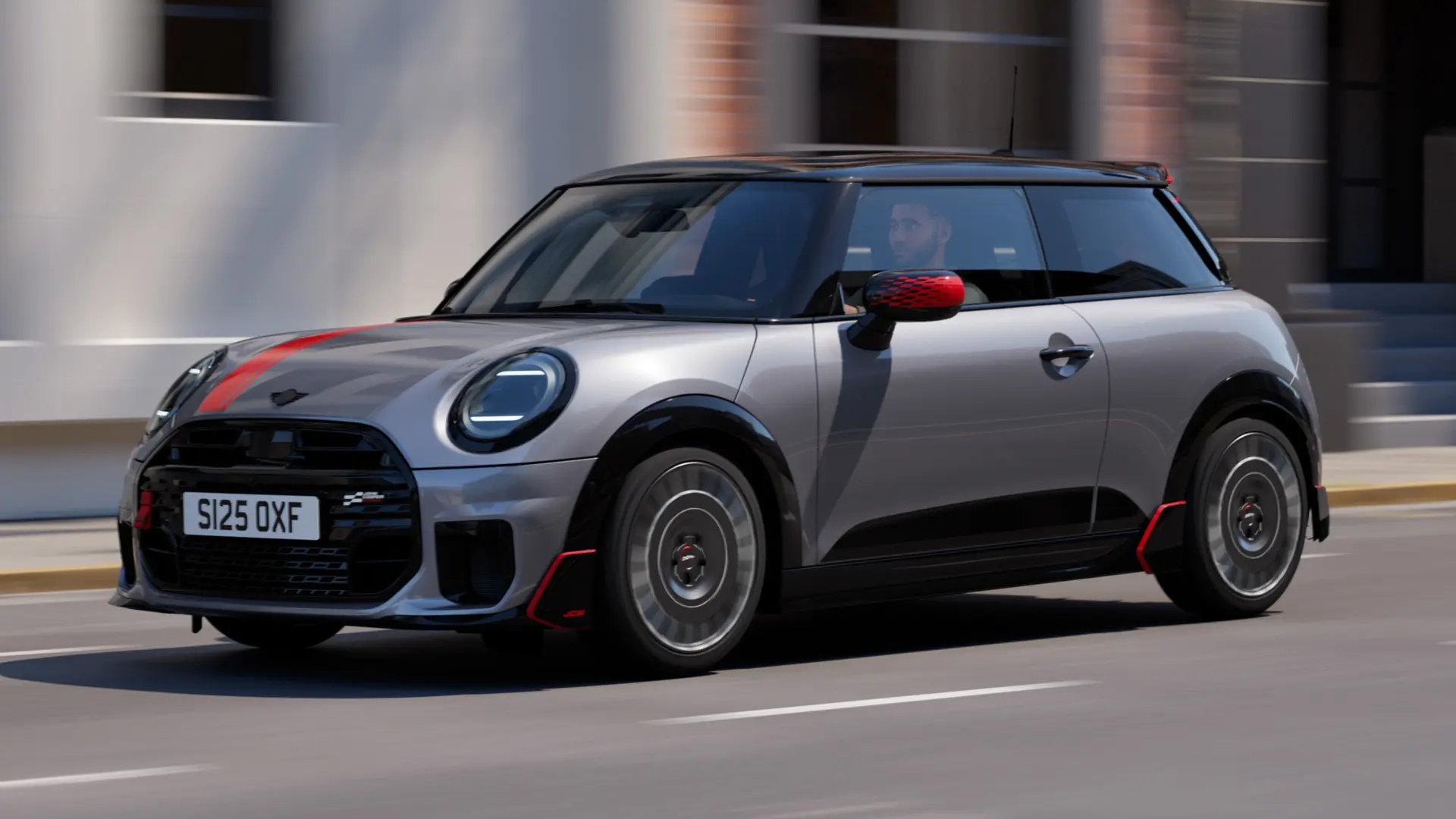 MINI Cooper s 3 vrati v barvi Melting Silver z deli John Cooper Works: pokrovi zunanjih ogledal in nalepko na pokrovu motorja v barvi Chili Red, strešnim spojlerjem in krilcem v črni barvi med vožnjo po mestni ulici – pogled od spredaj.